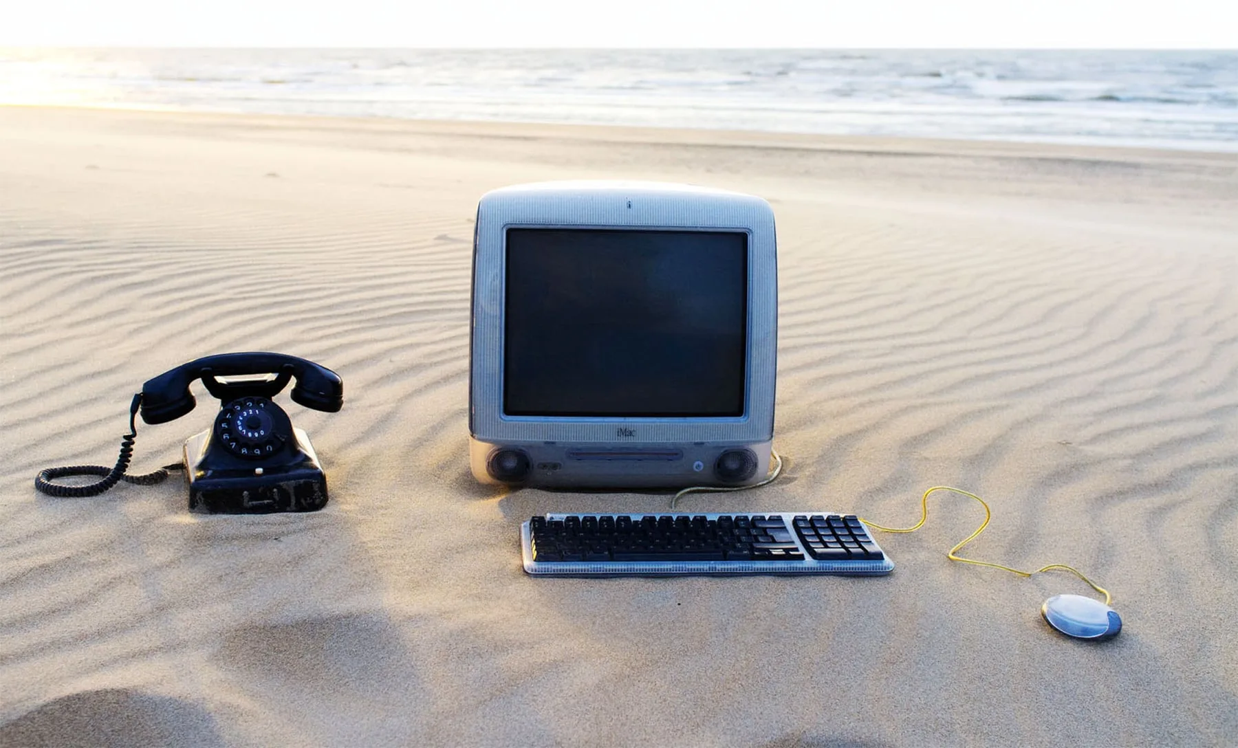 Rotary phone, iMac, keyboard and mouse arranged on a beach
