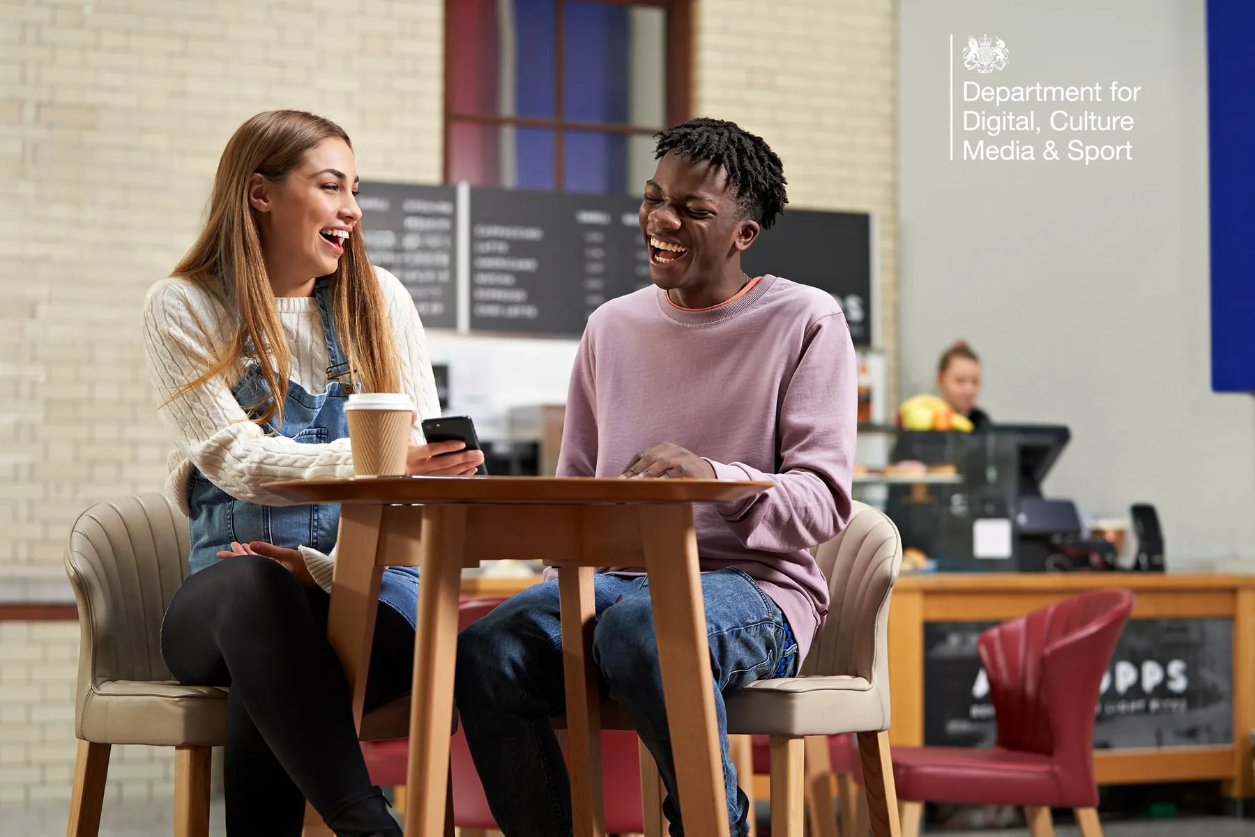 Two students laughing over a phone in a cafe, DCMS campaign