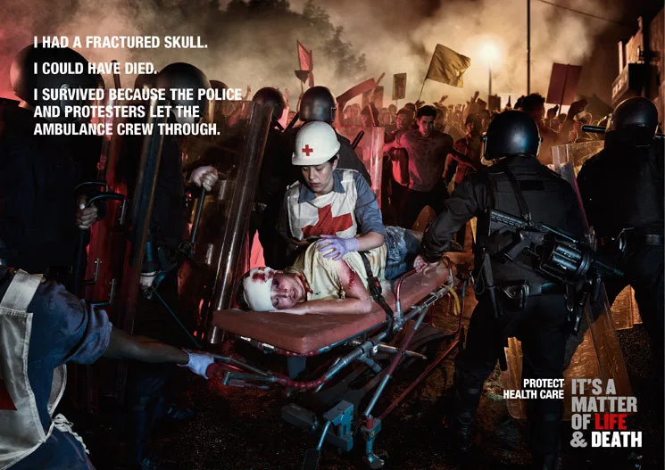 A red-cross medic tending an injured man on a stretcher between riot police and protesters — ICRC 'Health Care in Danger' campaign