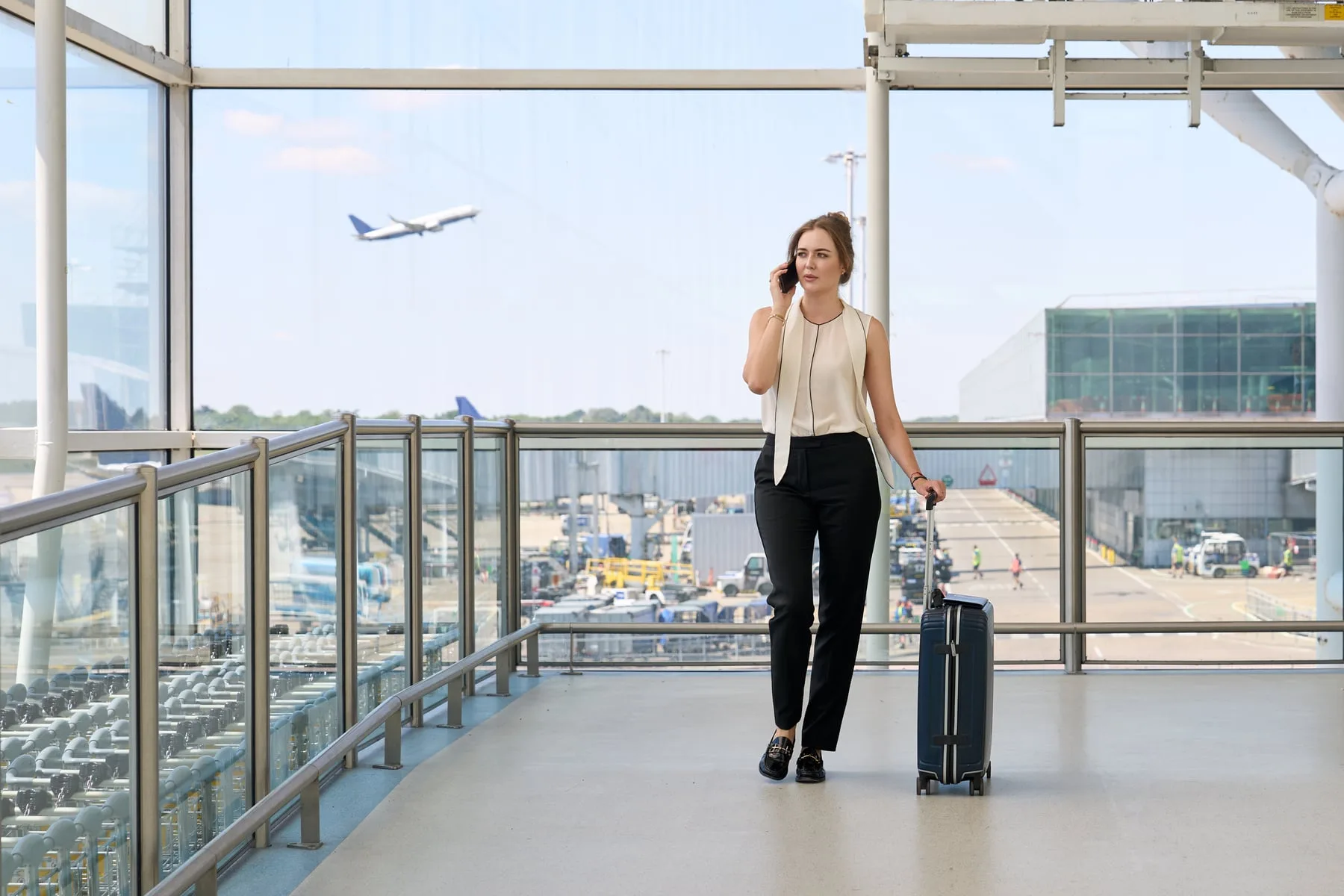 A business traveller on her phone with a suitcase at London Stansted Airport, a jet taking off behind — International SOS