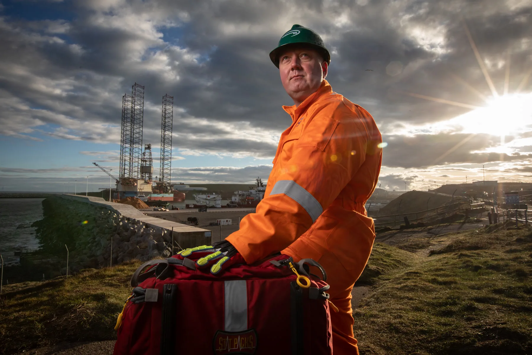 An International SOS medic in orange coveralls and hard hat with a first-response bag at the harbour at sunrise, oil rigs behind, Aberdeen
