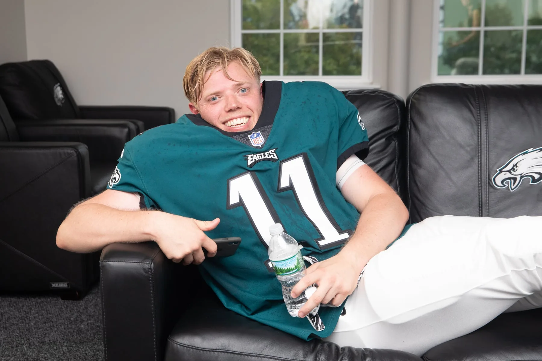 Rob Beckett lounging on a black Eagles sofa with a water bottle and phone — Sky 'Rob & Romesh Vs'