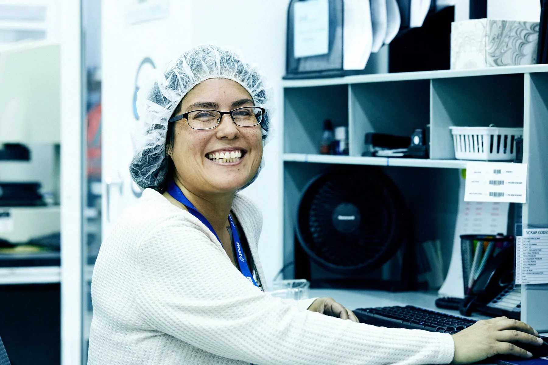 A West Pharmaceuticals colleague in a hairnet and branded lanyard laughing at her desk on the production floor, Arizona